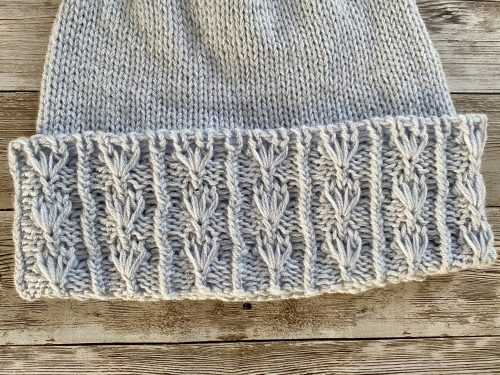 An image of a knitted Ginkgo Biloba hat in a soft ice blue color. The hat features a textured brim with a pattern resembling the fan-shaped leaves of the Ginkgo tree, adding an elegant and natural touch to the design. The background is a rustic wooden surface, enhancing the cozy and handmade aesthetic of the hat