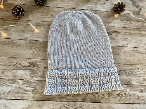 An image of a knitted Ginkgo Biloba hat in a soft ice blue color. The hat features a textured brim with a pattern resembling the fan-shaped leaves of the Ginkgo tree, adding an elegant and natural touch to the design. The background is a rustic wooden surface, enhancing the cozy and handmade aesthetic of the hat