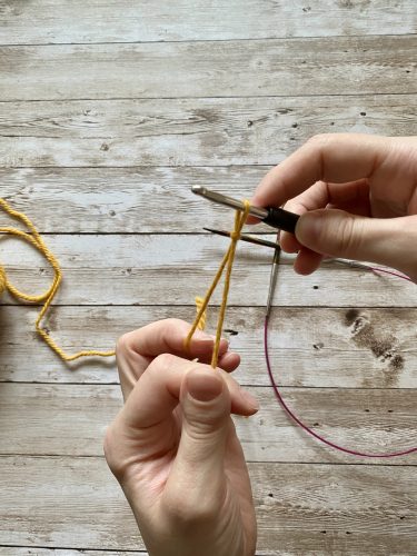 Demonstrating how to work the provisional cast on with white and yellow yarn, a blue chrochet hook and wooden needles on a wooden surface