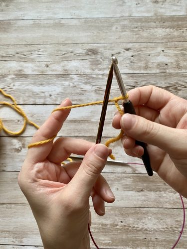 Demonstrating how to work the provisional cast on with white and yellow yarn, a blue chrochet hook and wooden needles on a wooden surface