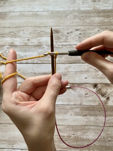 Demonstrating how to work the provisional cast on with white and yellow yarn, a blue chrochet hook and wooden needles on a wooden surface