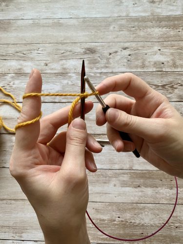 Demonstrating how to work the provisional cast on with white and yellow yarn, a blue chrochet hook and wooden needles on a wooden surface