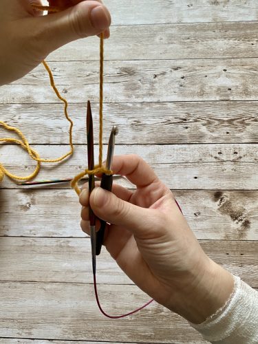 Demonstrating how to work the provisional cast on with white and yellow yarn, a blue chrochet hook and wooden needles on a wooden surface
