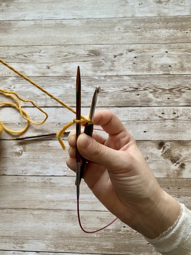 Demonstrating how to work the provisional cast on with white and yellow yarn, a blue chrochet hook and wooden needles on a wooden surface