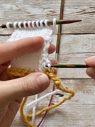 Demonstrating how to work the provisional cast on with white and yellow yarn, a blue chrochet hook and wooden needles on a wooden surface
