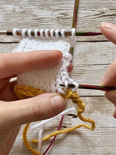 Demonstrating how to work the provisional cast on with white and yellow yarn, a blue chrochet hook and wooden needles on a wooden surface