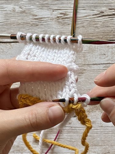 Demonstrating how to work the provisional cast on with white and yellow yarn, a blue chrochet hook and wooden needles on a wooden surface