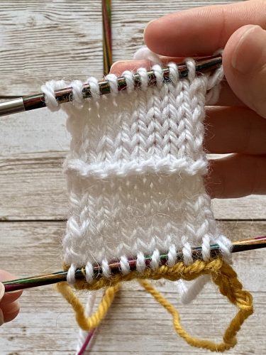 Demonstrating how to work the provisional cast on with white and yellow yarn, a blue chrochet hook and wooden needles on a wooden surface