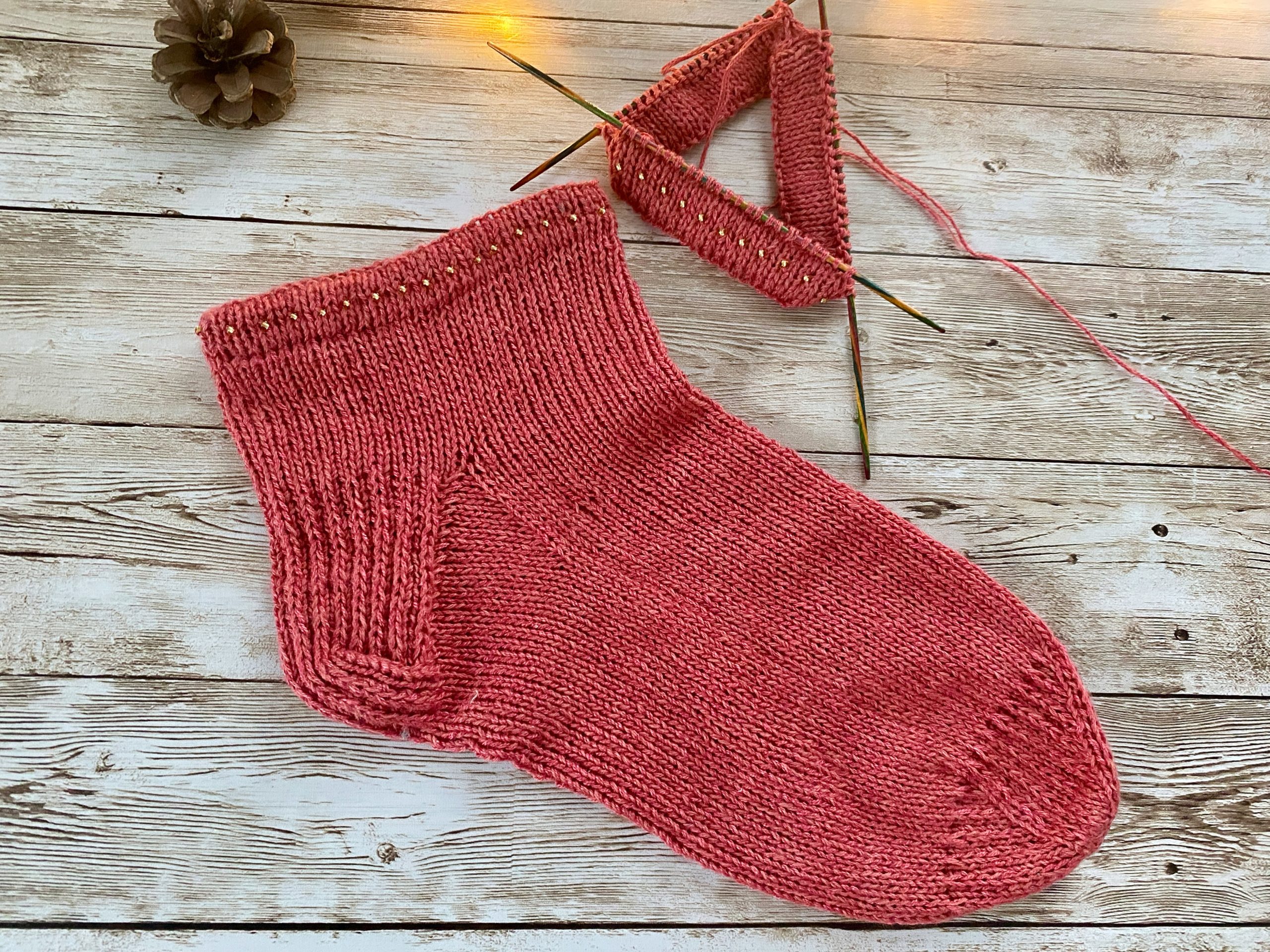A hand-knitted coral pink sock with ribbed cuff lies on a weathered wooden surface, next to knitting needles holding the beginning of a second matching sock. A small pinecone decorates the corner of the rustic backdrop, creating a cozy, crafty scene.