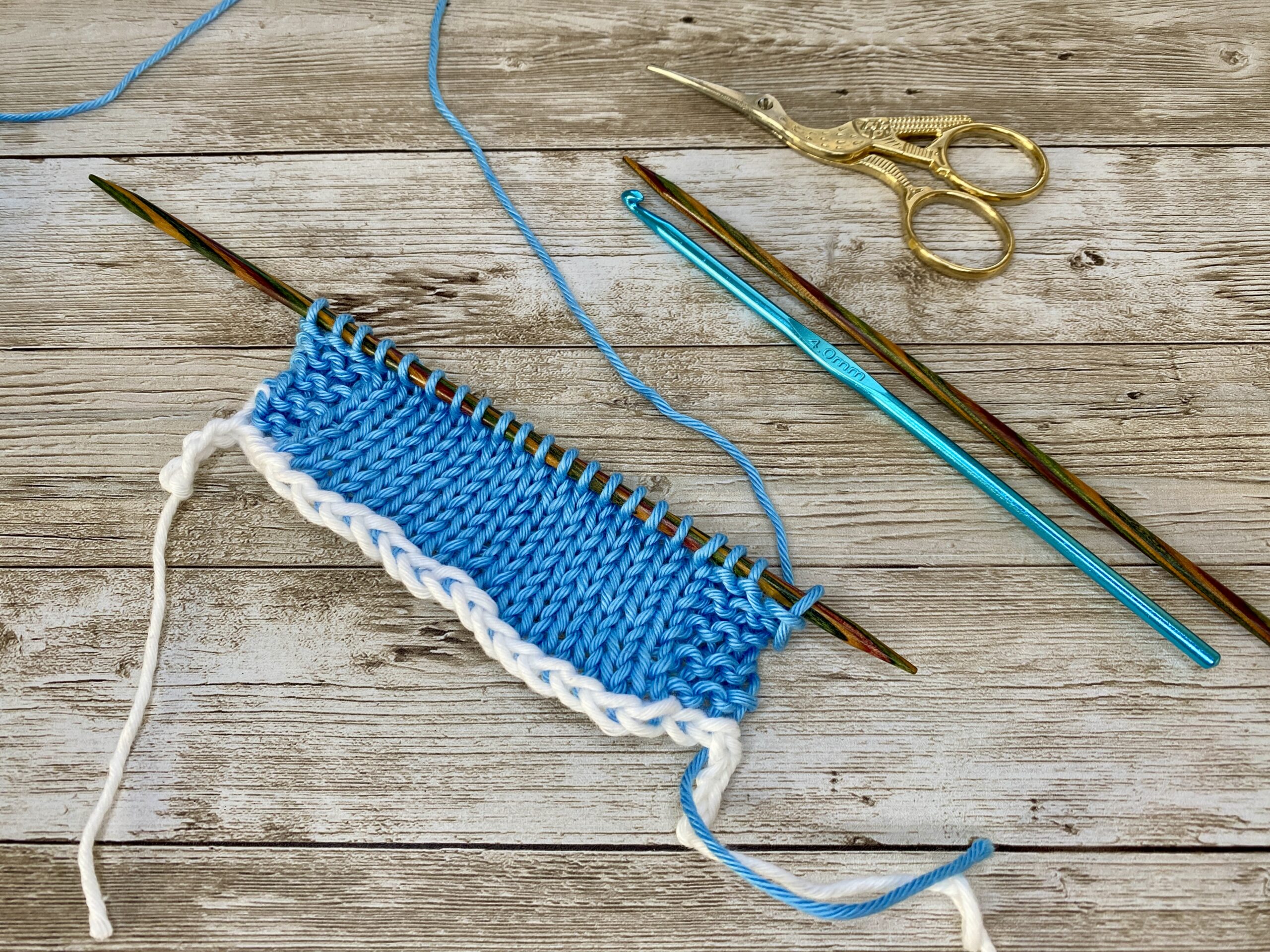 Demonstration of the provisional cast on knitting technique. A bright blue knitted swatch with a white crochet chain visible at the bottom edge is shown on wooden knitting needles. A crochet hook, knitting needle, and gold scissors are arranged nearby on a rustic wooden surface.