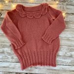 An image of the Little Ithilien sweater, a hand-knitted garment in a warm terracotta color. The sweater features a delicate scalloped neckline, long sleeves with textured cuffs, and a beautifully detailed hem with intricate patterns. The background is a rustic wooden surface adorned with soft, glowing string lights, adding a cozy and charming atmosphere to the presentation.