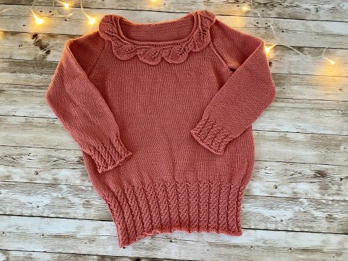 An image of the Little Ithilien sweater, a hand-knitted garment in a warm terracotta color. The sweater features a delicate scalloped neckline, long sleeves with textured cuffs, and a beautifully detailed hem with intricate patterns. The background is a rustic wooden surface adorned with soft, glowing string lights, adding a cozy and charming atmosphere to the presentation.