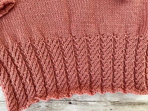 An image of the Little Ithilien sweater, a hand-knitted garment in a warm terracotta color. The sweater features a delicate scalloped neckline, long sleeves with textured cuffs, and a beautifully detailed hem with intricate patterns. The background is a rustic wooden surface adorned with soft, glowing string lights, adding a cozy and charming atmosphere to the presentation.