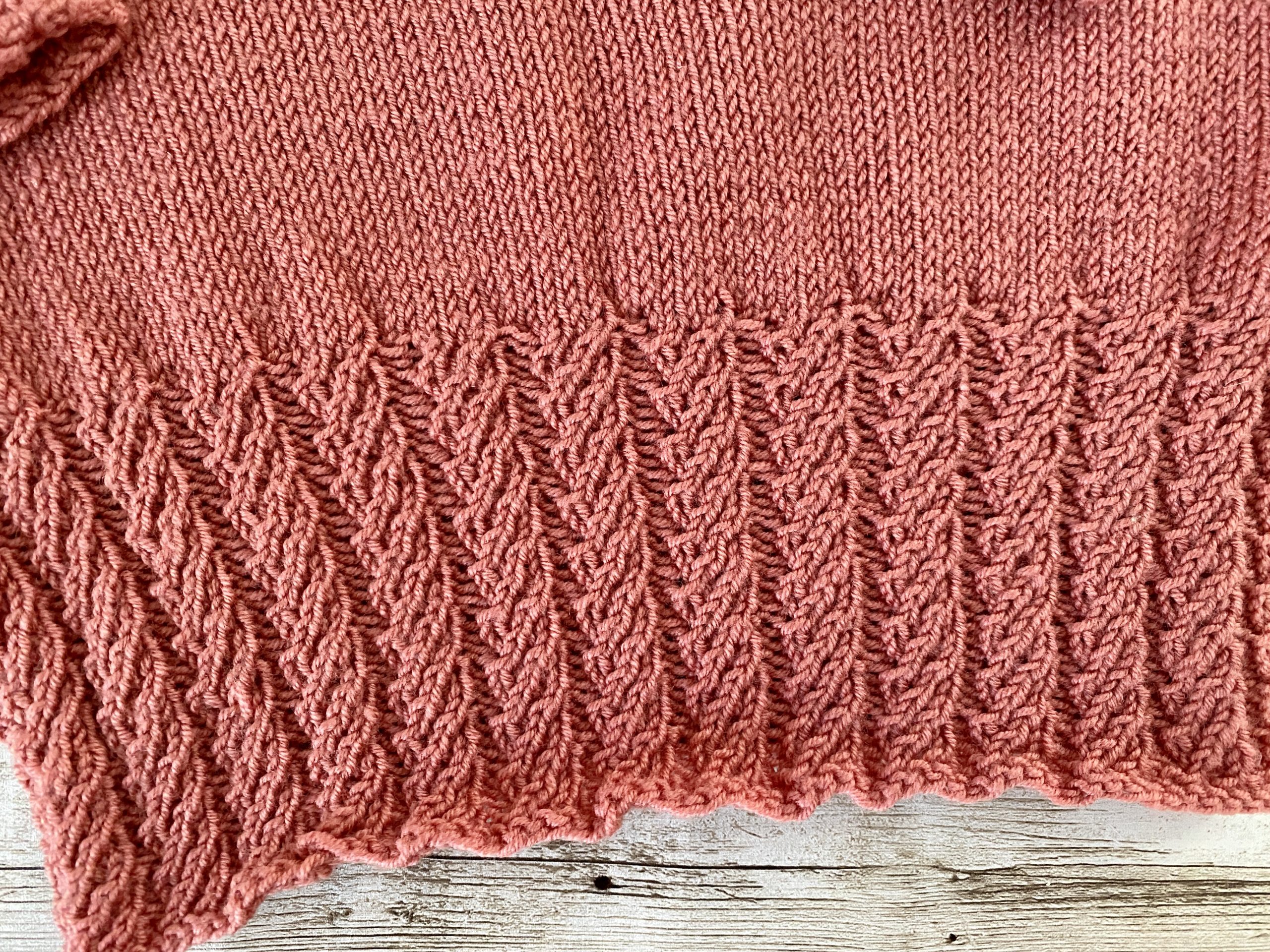 An image of the Little Ithilien sweater, a hand-knitted garment in a warm terracotta color. The sweater features a delicate scalloped neckline, long sleeves with textured cuffs, and a beautifully detailed hem with intricate patterns. The background is a rustic wooden surface adorned with soft, glowing string lights, adding a cozy and charming atmosphere to the presentation.