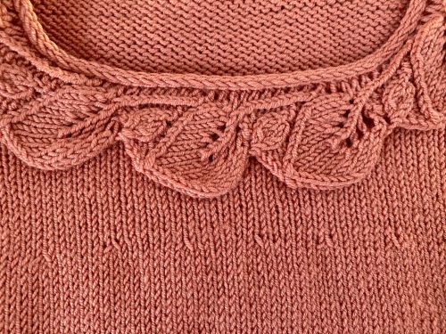 An image of the Little Ithilien sweater, a hand-knitted garment in a warm terracotta color. The sweater features a delicate scalloped neckline, long sleeves with textured cuffs, and a beautifully detailed hem with intricate patterns. The background is a rustic wooden surface adorned with soft, glowing string lights, adding a cozy and charming atmosphere to the presentation.