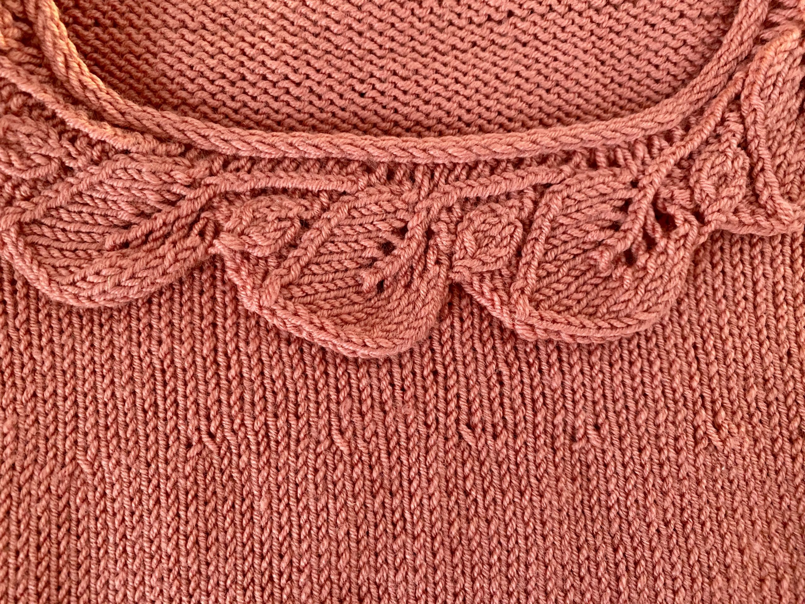 An image of the Little Ithilien sweater, a hand-knitted garment in a warm terracotta color. The sweater features a delicate scalloped neckline, long sleeves with textured cuffs, and a beautifully detailed hem with intricate patterns. The background is a rustic wooden surface adorned with soft, glowing string lights, adding a cozy and charming atmosphere to the presentation.