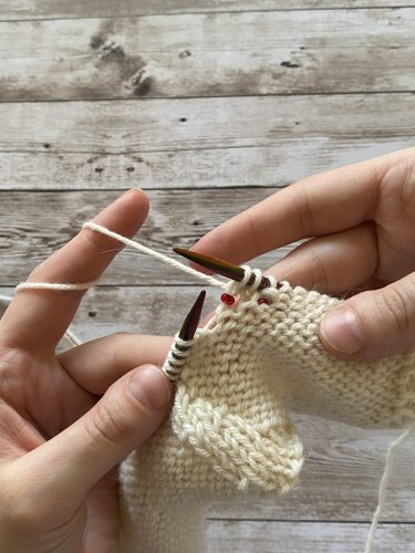 Step-by-step tutorial image demonstrating one of three techniques for threading beads onto yarn for knitting: using a hook, dental floss or thread, or pre-threading. The image shows the tools, materials, and process clearly to guide the viewer through the technique.