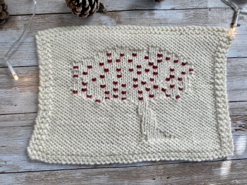 A knitted swatch featuring a textured tree design with red beads accenting the tree's canopy. The swatch is made with cream-colored yarn and bordered with a garter stitch edge. The background includes a rustic wooden surface, pinecones, and soft string lights, adding a cozy and festive atmosphere.