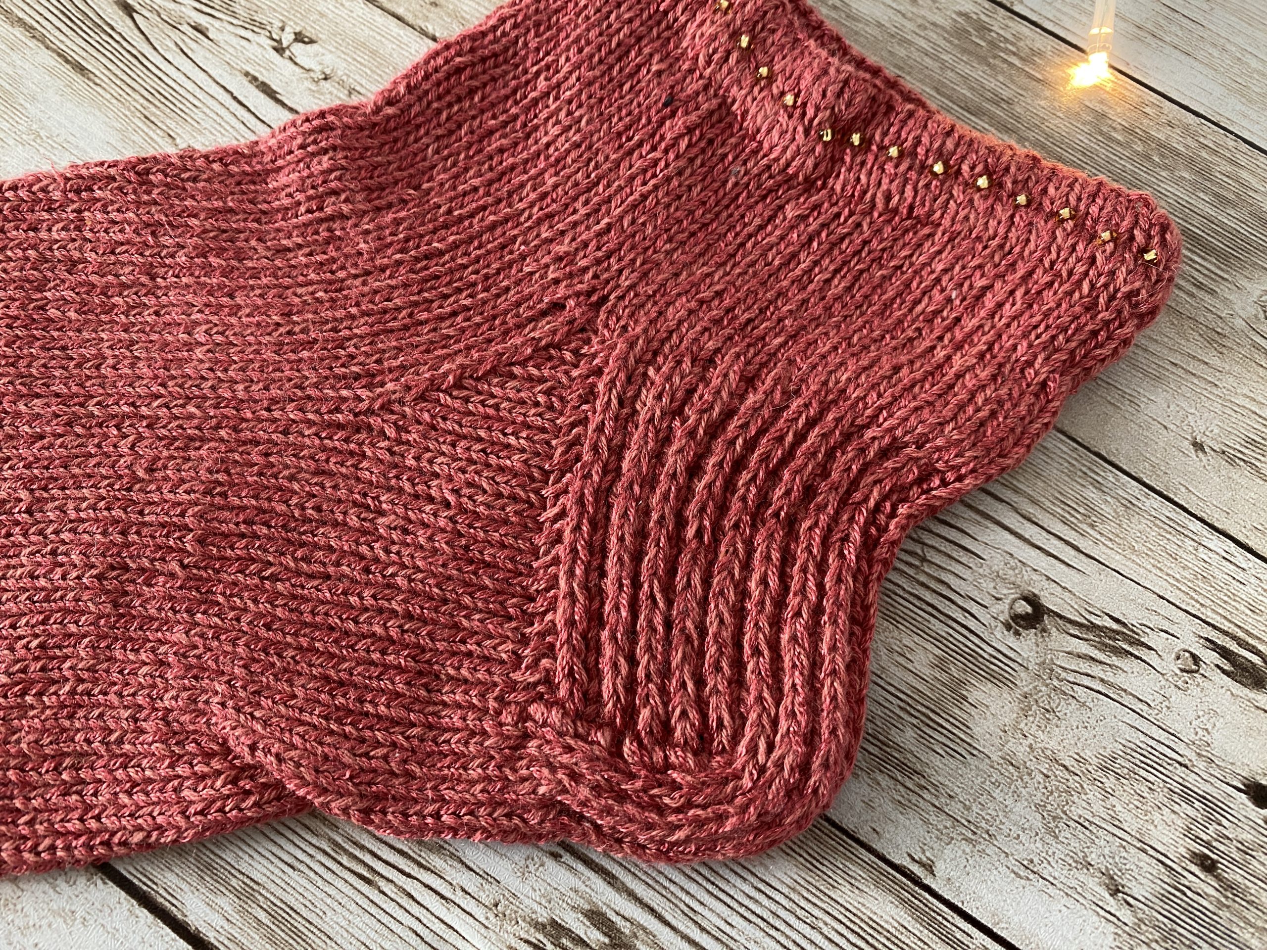 A hand-knitted sock in a warm pink color, displayed on a rustic wooden surface. The completed sock features a ribbed cuff, a reinforced heel, and a smooth stockinette body. Nearby, a second sock is in progress on double-pointed needles, with yarn and tools arranged neatly. A pinecone and soft string lights add a cozy touch to the scene.