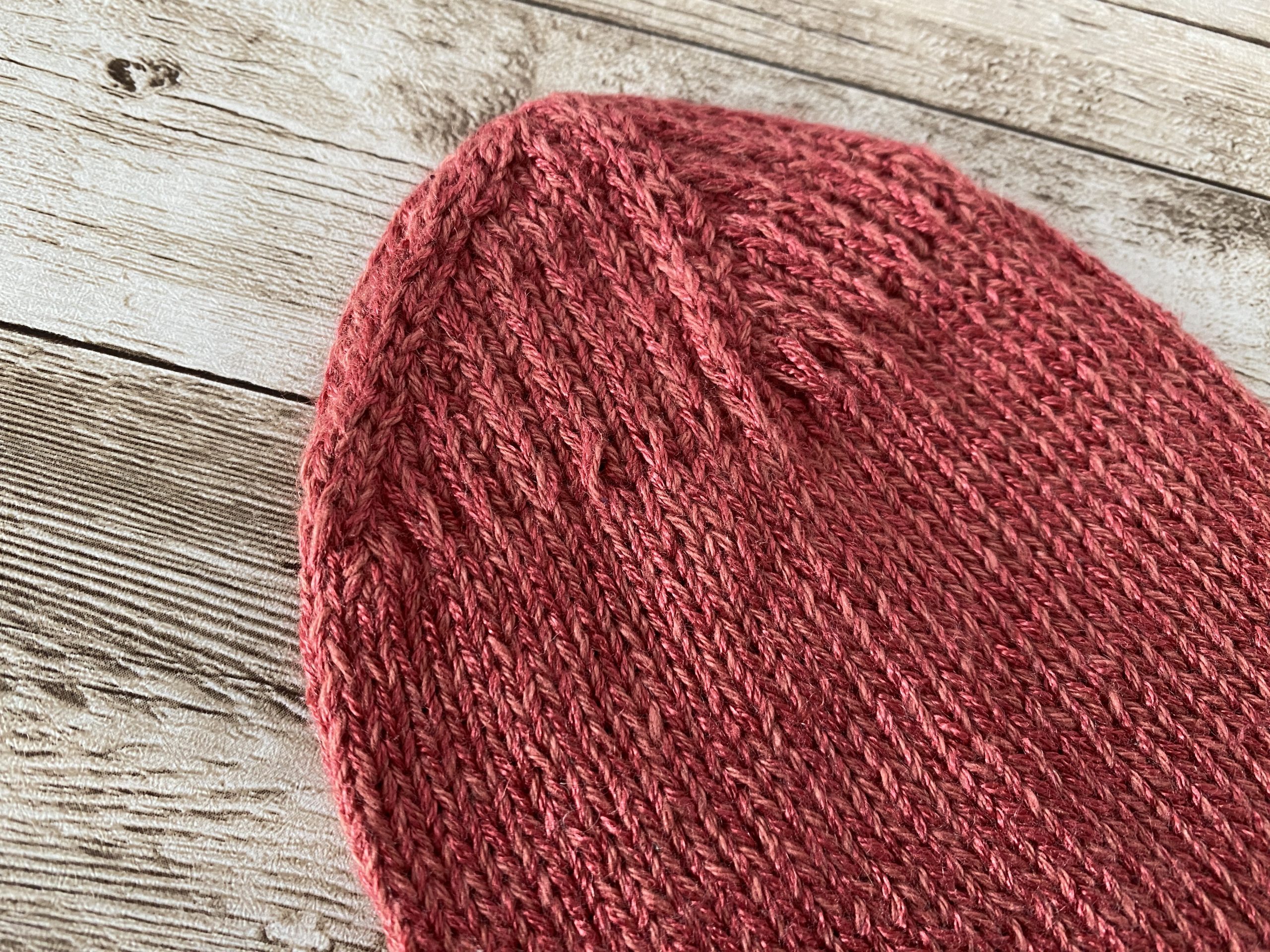A hand-knitted sock in a warm pink color, displayed on a rustic wooden surface. The completed sock features a ribbed cuff, a reinforced heel, and a smooth stockinette body. Nearby, a second sock is in progress on double-pointed needles, with yarn and tools arranged neatly. A pinecone and soft string lights add a cozy touch to the scene.