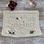 Cream-colored knitted swatch laid on a wooden surface, featuring textured stitching with floral embroidery details in green and white, accented by decorative lights and red flowers in the background.