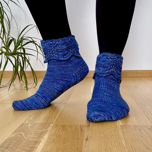 A pair of hand-knitted blue socks with a textured, lacy cuff featuring decorative buttons. The socks are made with variegated blue yarn, showcasing subtle color variations, and are displayed on a rustic wooden surface.