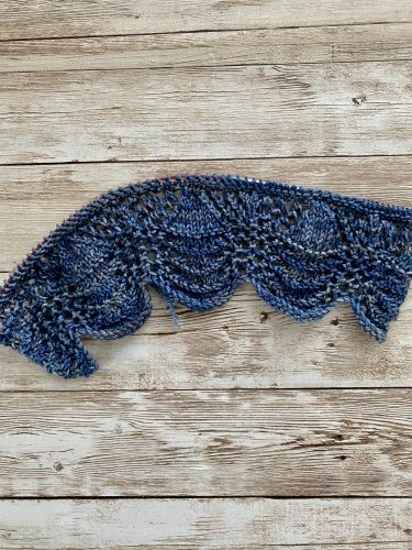 A pair of hand-knitted blue socks with a textured, lacy cuff featuring decorative buttons. The socks are made with variegated blue yarn, showcasing subtle color variations, and are displayed on a rustic wooden surface.