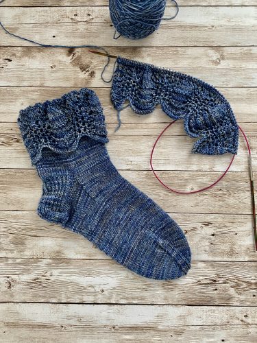 A pair of hand-knitted blue socks with a textured, lacy cuff featuring decorative buttons. The socks are made with variegated blue yarn, showcasing subtle color variations, and are displayed on a rustic wooden surface.