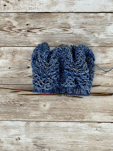 A pair of hand-knitted blue socks with a textured, lacy cuff featuring decorative buttons. The socks are made with variegated blue yarn, showcasing subtle color variations, and are displayed on a rustic wooden surface.