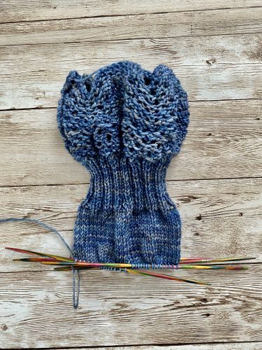 A pair of hand-knitted blue socks with a textured, lacy cuff featuring decorative buttons. The socks are made with variegated blue yarn, showcasing subtle color variations, and are displayed on a rustic wooden surface.