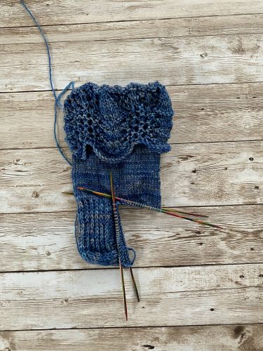 A pair of hand-knitted blue socks with a textured, lacy cuff featuring decorative buttons. The socks are made with variegated blue yarn, showcasing subtle color variations, and are displayed on a rustic wooden surface.