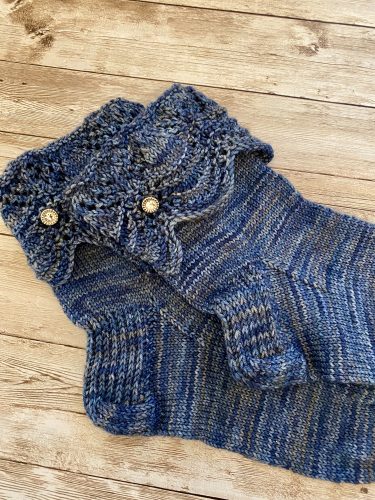 A pair of hand-knitted blue socks with a textured, lacy cuff featuring decorative buttons. The socks are made with variegated blue yarn, showcasing subtle color variations, and are displayed on a rustic wooden surface.