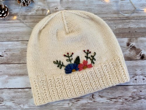 A hand-knitted cream-colored hat with a ribbed brim and a smooth stockinette body, adorned with delicate embroidered flowers in blue, pink, and red, along with green leaves. The hat is displayed on a rustic wooden surface, surrounded by pinecones and soft string lights, creating a cozy and charming atmosphere.