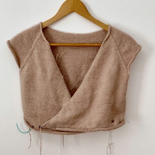 A partially completed Robinson Wrap Cardigan in a soft beige color, displayed on a wooden hanger against a white wall. The cardigan features a wrap-style front with short sleeves and visible unfinished edges, with loose yarn strands hanging from the bottom.
