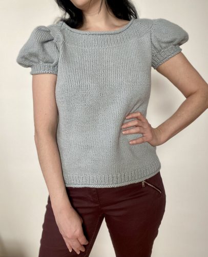 A woman wearing the Anne Sweater in light gray, showcasing its puffed short sleeves and fitted silhouette. The sweater is paired with burgundy pants, highlighting its elegant and versatile design.
