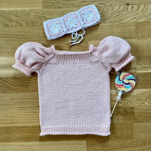A soft pink Anne Sweater with puffed short sleeves, displayed on a wooden surface. Accompanying the sweater are a crocheted headband with daisy motifs and a colorful lollipop, creating a playful and whimsical arrangement.