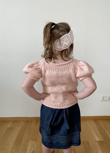 A child wearing the Anne Sweater in soft pink, viewed from the back. The sweater features puffed short sleeves and a snug fit, paired with a denim skirt and a crocheted headband with daisy motifs.