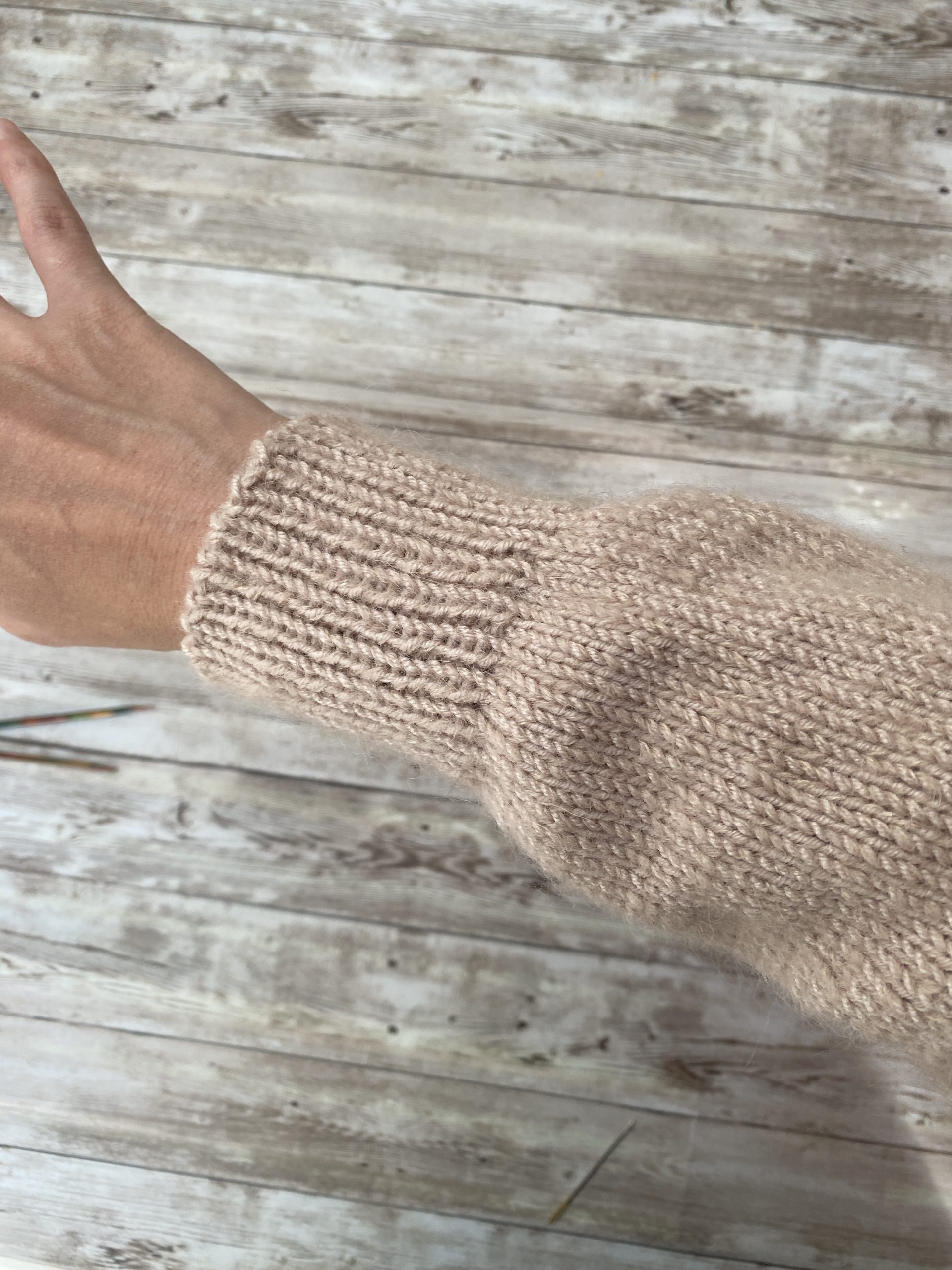 A close-up of a sleeve from the Robinson Wrap Cardigan, showcasing the soft beige knit fabric. The sleeve features a simple stockinette stitch with a snug ribbed cuff, worn on an outstretched arm against a rustic wooden background.