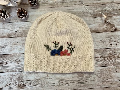 A hand-knitted cream-colored hat with a ribbed brim and a smooth stockinette body, adorned with delicate embroidered flowers in blue, pink, and red, along with green leaves. The hat is displayed on a rustic wooden surface, surrounded by pinecones and soft string lights, creating a cozy and charming atmosphere.