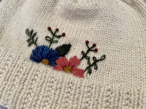 A hand-knitted cream-colored hat with a ribbed brim and a smooth stockinette body, adorned with delicate embroidered flowers in blue, pink, and red, along with green leaves. The hat is displayed on a rustic wooden surface, surrounded by pinecones and soft string lights, creating a cozy and charming atmosphere.