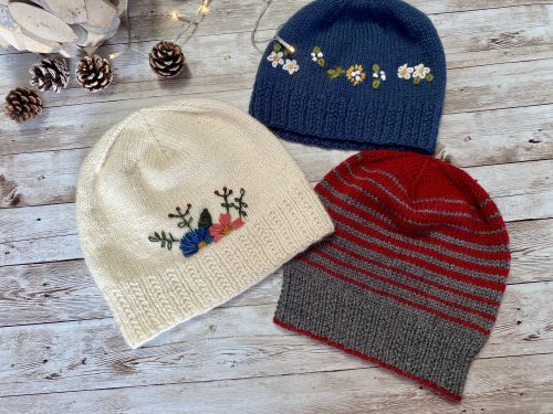 Three knitted Williams hats are displayed on a wooden surface with small pinecones scattered around. The hats include a white one with colorful floral embroidery, a blue one with white and yellow flower embroidery, and a red and gray striped one. The cozy setup evokes a warm, handmade feel.