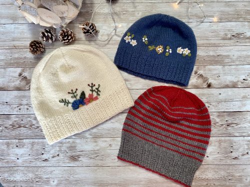 Three hand-knitted hats with a ribbed brim and a smooth stockinette body, adorned with delicate embroidered flowers in blue, pink, and red, along with green leaves. The hat is displayed on a rustic wooden surface, surrounded by pinecones and soft string lights, creating a cozy and charming atmosphere.