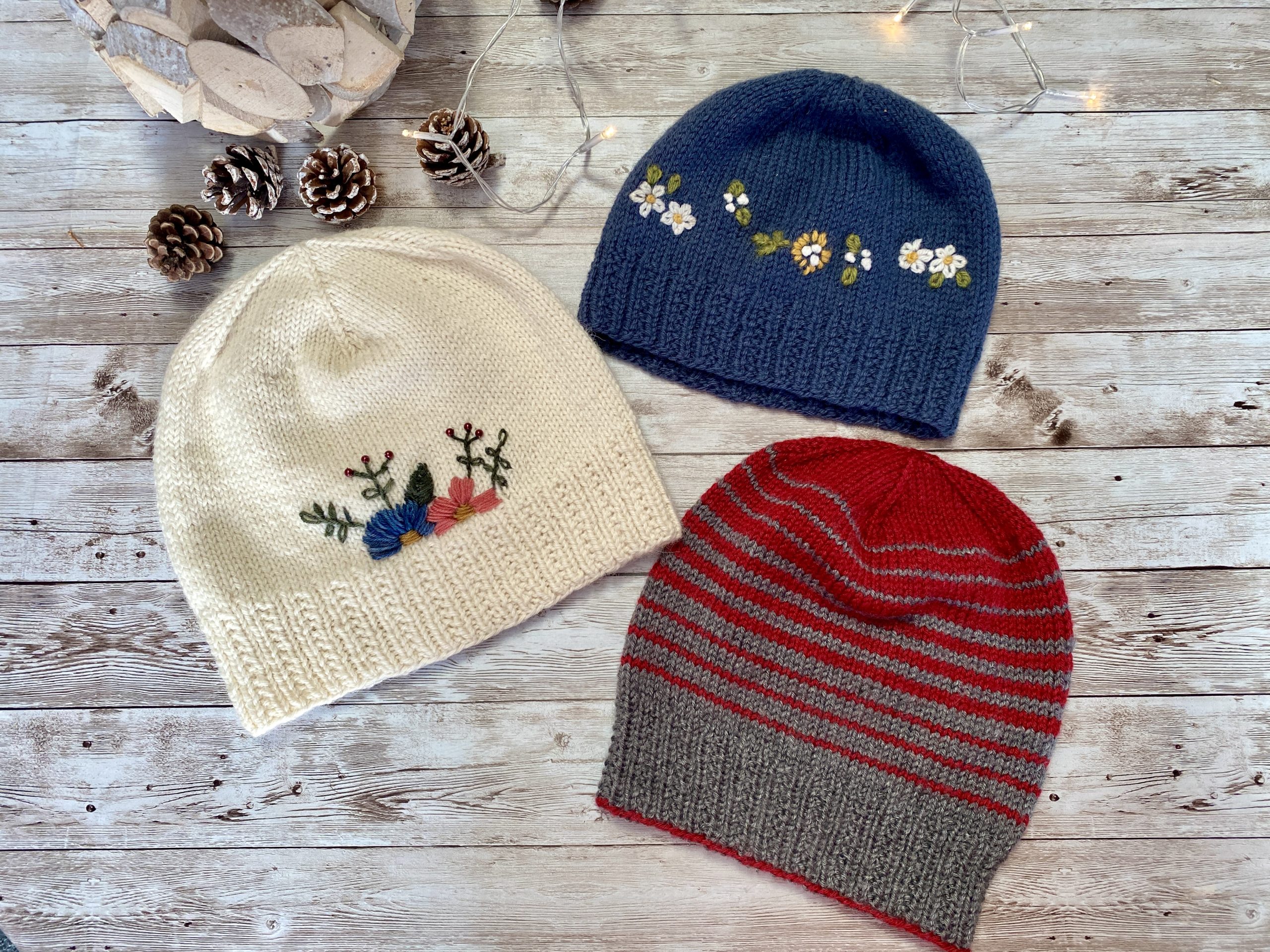 Three hand-knitted hats with a ribbed brim and a smooth stockinette body, adorned with delicate embroidered flowers in blue, pink, and red, along with green leaves. The hat is displayed on a rustic wooden surface, surrounded by pinecones and soft string lights, creating a cozy and charming atmosphere.