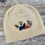 A hand-knitted cream-colored hat with a ribbed brim and a smooth stockinette body, adorned with delicate embroidered flowers in blue, pink, and red, along with green leaves. The hat is displayed on a rustic wooden surface, surrounded by pinecones and soft string lights, creating a cozy and charming atmosphere.