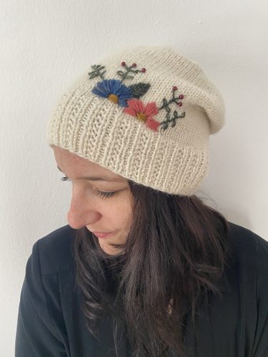 Me wearing the Williams hat - cream-colored - with a ribbed brim and a smooth stockinette body, adorned with delicate embroidered flowers in blue, pink, and red, along with green leaves. The hat is displayed on a rustic wooden surface, surrounded by pinecones and soft string lights, creating a cozy and charming atmosphere.