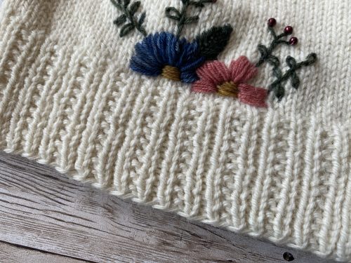 A hand-knitted cream-colored hat with a ribbed brim and a smooth stockinette body, adorned with delicate embroidered flowers in blue, pink, and red, along with green leaves. The hat is displayed on a rustic wooden surface, surrounded by pinecones and soft string lights, creating a cozy and charming atmosphere.