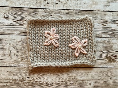 A knitted swatch from the Fluffy Daisy Cardigan, designed by TheKnitStitch, displayed on a rustic wooden surface. The swatch is made with chunky beige yarn in a stockinette stitch and features two embroidered daisies in soft pink yarn with cream-colored centers, showcasing the decorative floral detail of the cardigan design.