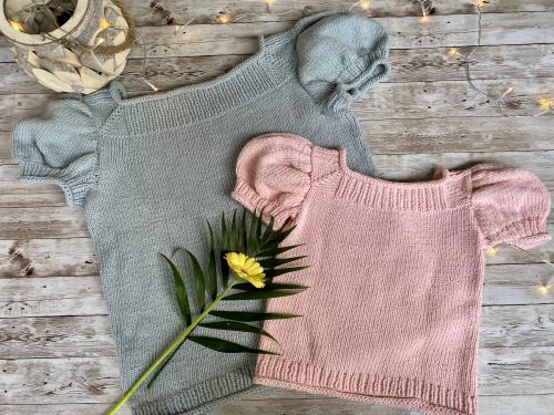 Two Anne Sweaters, one in light gray and the other in soft pink, laid flat on a rustic wooden surface. Both feature puffed short sleeves and a simple stockinette stitch design. A yellow flower and decorative lights add a charming touch to the display.