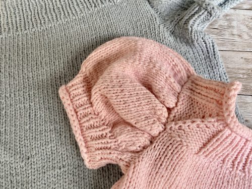 A close-up of the Anne Sweater in light gray and soft pink, focusing on the puffed short sleeves and the smooth stockinette stitch texture. The two sweaters are layered together on a rustic wooden surface, highlighting their delicate craftsmanship.