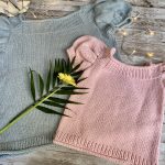 Two Anne Sweaters, one in light gray and the other in soft pink, laid flat on a rustic wooden surface. Both feature puffed short sleeves and a simple stockinette stitch design. A yellow flower and decorative lights add a charming touch to the display.
